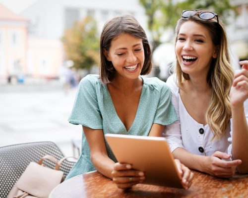 Holidays, tourism, technology friends concept. Beautiful girls looking at tablet pc computer in cafe outside