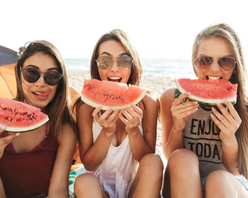 Cheerful happy friends camping at the beach, eating watermelon, laughing